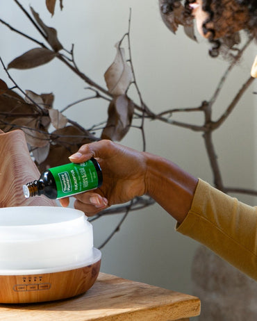 Person adding essential oil to a diffuser on a wooden table with decorative branches in the background.