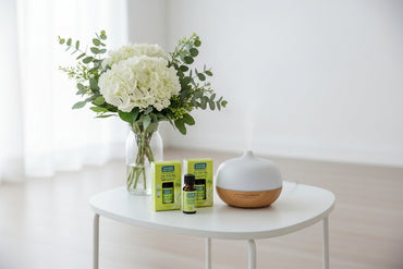Aromatherapy diffuser and essential oils on a white table with a vase of flowers in the background.